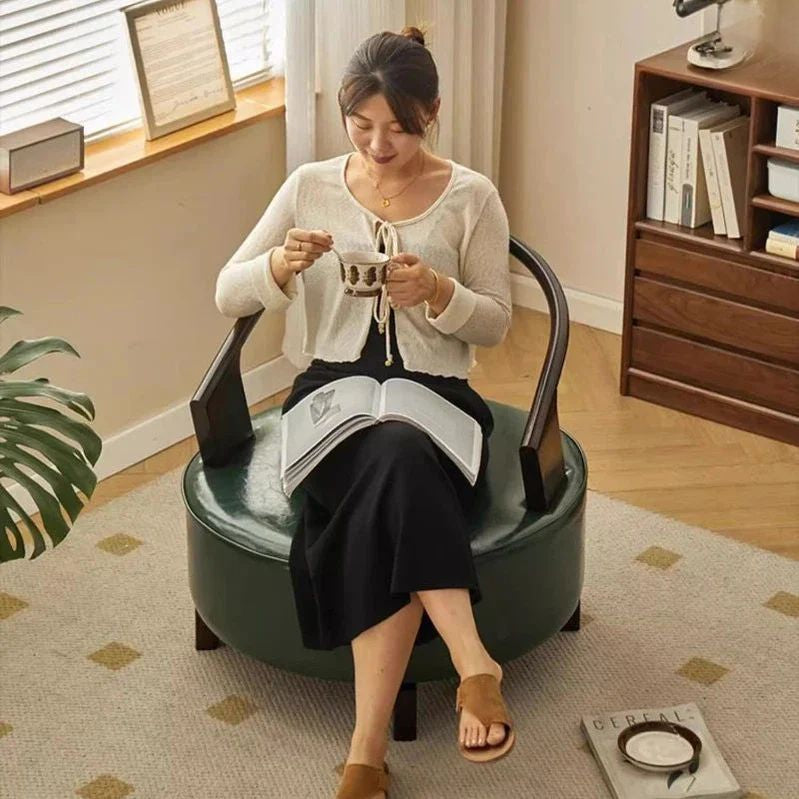 "Woman sitting on a green chair reading a book and drinking from a cup in a cozy room."