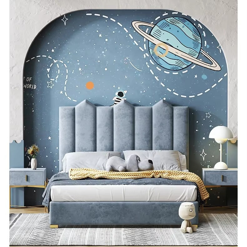 "Children's bedroom featuring a modern bed with a blue upholstered headboard, placed against a wall with a space-themed mural, including a planet and stars."
