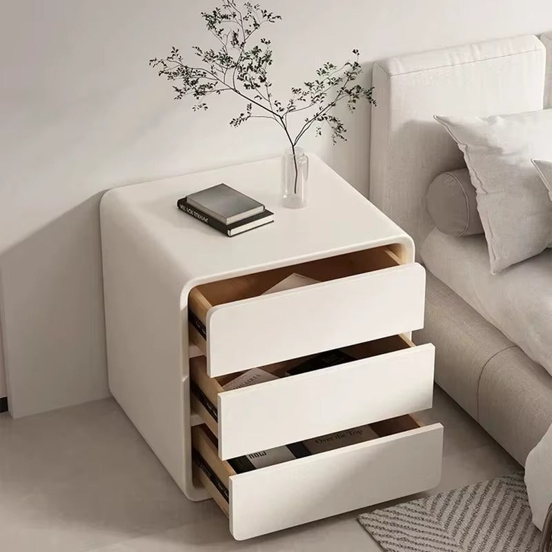 "White nightstand with three drawers next to a bed in a bedroom setting."