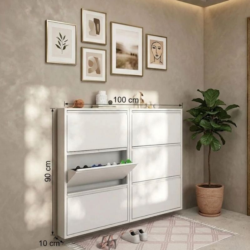 White shoe cabinet with measurements against a beige wall with framed art and a plant.